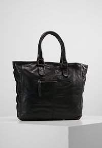 Sac cabas en cuir noir avec deux poignées, une poche avant zippée et une texture lisse. Présente des côtés légèrement froncés pour la forme.