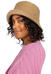 Beige bøttehat lavet af tekstureret stof, med en bred skygge og et lille metal-logo accent på siden, båret af en person i en pink top.