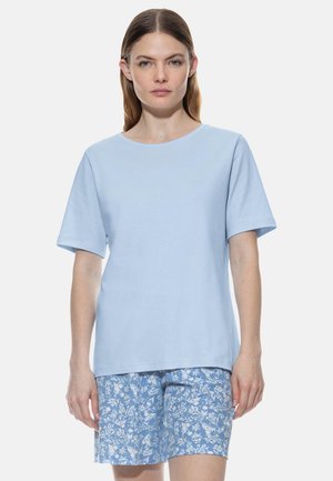 Vrouw met steil bruin haar die een lichtblauw T-shirt met korte mouwen en blauwe korte broek met bloemenpatroon draagt, staand tegen een effen achtergrond.