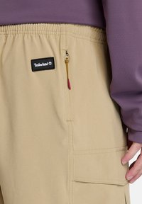 Béžové cargo šortky s elastickým pasem, boční kapsou na zip a nášivkou Timberland; spárované s fialovým topem s dlouhým rukávem.