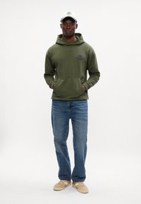 Olivovo zelená mikina s klokankou a čiernym logom, v kombinácii s modrými džínsami a béžovými teniskami. Čiapka má svetložltý šilt a logo.