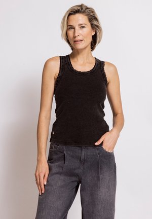 Woman with short blonde hair wears a sleeveless black top and high-waisted dark gray pants, standing against a plain light background.