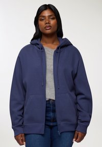 Donna che indossa un hoodie blu navy con zip e lacci sopra una maglietta grigia e jeans blu, in piedi contro uno sfondo chiaro e semplice.