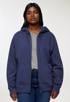 Donna che indossa un hoodie blu navy con zip e lacci sopra una maglietta grigia e jeans blu, in piedi contro uno sfondo chiaro e semplice.