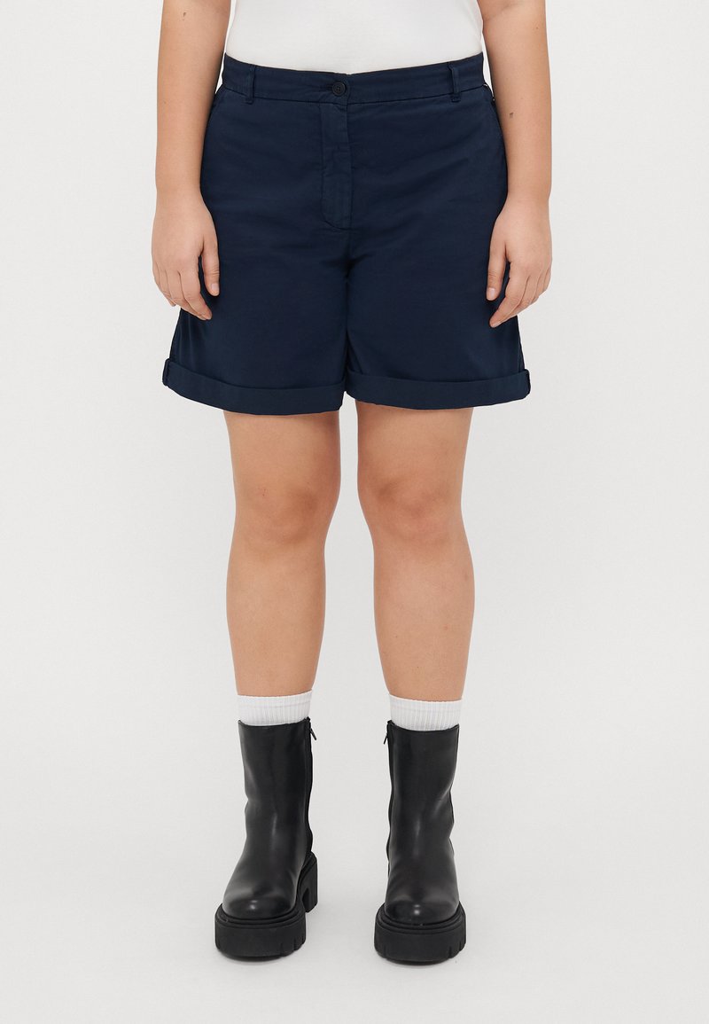 Mørkeblå shorts med oppbrett, laget av glatt stoff. Kombinert med svarte ankelstøvler og hvite sokker. Enkel og funksjonell design.