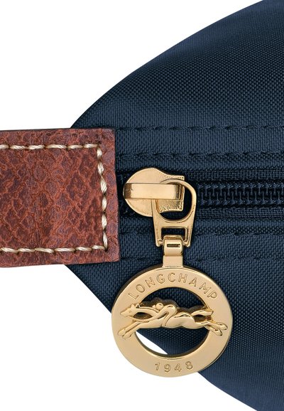 Navy nylon bag detail featuring a gold zipper and charm with a horse design, finished with a brown leather accent and stitched details.
