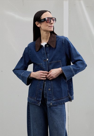 Veste en denim avec un col en velours côtelé marron, boutons sur le devant et deux poches poitrine ; assortie d'un pantalon en denim. Le cadre présente des lunettes de soleil.