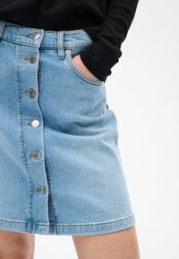 En person som bär en ljusblå knapp-fram denimkjol med handen i fickan och en svart långärmad topp mot en vit bakgrund.