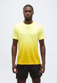 Άνδρας με αθλητική φανέλα με κίτρινη διαβάθμιση και κοντά μαύρα σορτς, στέκεται μπροστά σε απλό φωτεινό φόντο.