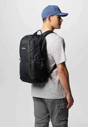 Giovane uomo che indossa un cappellino blu, una maglietta grigia chiara, pantaloni cargo grigi, con uno zaino nero Columbia, rivolto di lato.