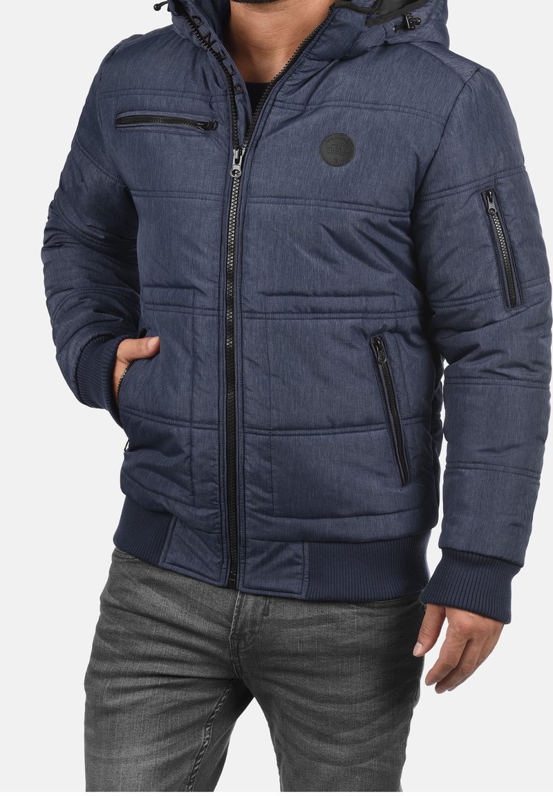 Homme portant une veste matelassée bleu marine avec plusieurs poches zippées et un jean gris, debout, une main dans la poche.