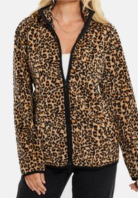 Kobieta nosząca polar z zamkiem błyskawicznym w print zwierzęcy, pod którym ma białą bluzkę i czarne spodnie, z złotym naszyjnikiem.