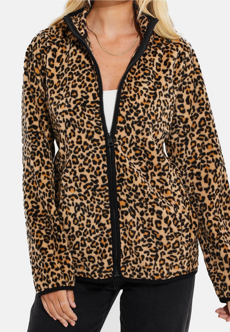 Kobieta nosząca polar z zamkiem błyskawicznym w print zwierzęcy, pod którym ma białą bluzkę i czarne spodnie, z złotym naszyjnikiem.