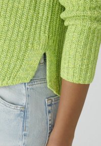 Nærbillede af en person iført en limegrøn ribstrikket sweater med en sideslids, parret med lyseblå denimjeans.