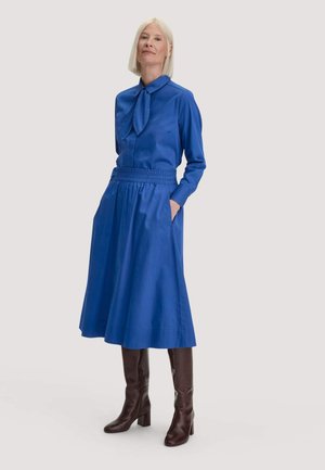 Frau mit kurzen blonden Haaren, die ein blaues langärmeliges Kleid mit Schlipskragen und kniehohe dunkelbraune Stiefel trägt, steht vor einem einfarbigen Hintergrund.