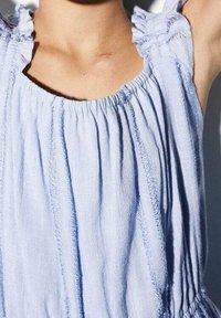 Vaaleansininen hihaton toppi, jossa on kerrostettua kangasta, rypytettyjä yksityiskohtia kaula-aukossa ja löysä, teksturoitu muotoilu.