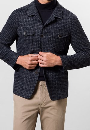 Mann, der eine dunkelblaue Hahnentrittmusterjacke über einen schwarzen Rollkragenpullover zuknöpft, beige Hosen trägt, vor schlichtem hellem Hintergrund.