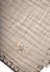 Gaišākas krēmkrāsas muslīna sega ar sakrustotu tekstūru, kurai ir izšūta mala un birka ar krāsainiem zvaigznēm un mēnesiem.