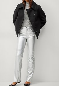 Manteau en laine noir à col rond, pull gris ajusté, pantalon en simili cuir argenté brillant, et chaussures à talons hauts noires.