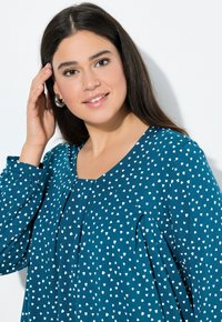 Jonge vrouw met lang, donker haar die haar gezicht aanraakt, met zilveren creolen en een teal blouse met witte abstracte stippen.