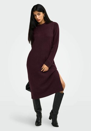 Femme aux longs cheveux foncés porte une robe-pull bordeaux à manches longues avec une fente sur le côté et des bottes noires hautes jusqu'au genou à talons, sur un fond uni.
