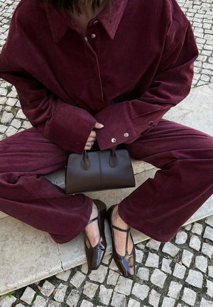 Conjunto de pana burdeos con mangas amplias y detalles en dorado, combinado con un bolso de cuero marrón y zapatos brillantes de punta marrón.