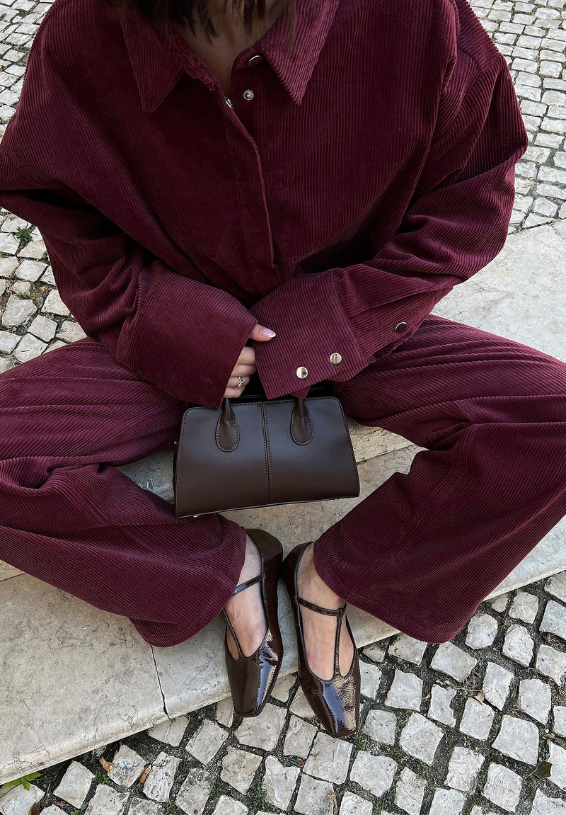 Completo in velluto a coste bordeaux con maniche ampie e hardware dorato, abbinato a una borsa in pelle marrone e scarpe lucide marroni con punta a punta.