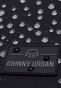 Fekete textil, amelyen kör alakú vízcseppek találhatók, egy kiemelkedő, szürke színű, "JOHNNY URBAN" feliratú címkével. Érdes felületi részletek láthatók.
