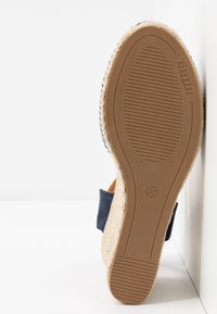 Semelle d’une sandale espadrille compensée avec semelle extérieure en caoutchouc, taille 37, bride bleu marine et semelle intérieure en jute tressée appuyée contre un mur blanc.