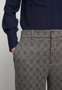 Detail ruky v kapse, na sobě šedé kostkované kalhoty s poutky na pásek a tmavě modrou dlouhou košili zasunutou vevnitř.