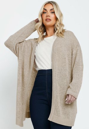 Beige Strickjacke aus strukturiertem Strickstoff, offener Front, lange Ärmel mit Rippbündchen, kombiniert mit einem hellen T-Shirt und dunklen Jeans.