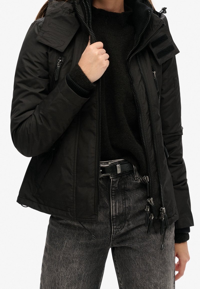 Personne portant une veste matelassée noire par-dessus un pull noir et un jean gris, une main tenant la fermeture éclair de la veste près de la poitrine.