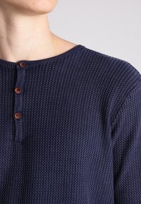 Pull en maille bleu marine avec un motif texturé, encolure arrondie et trois boutons marron sur la patte de boutonnage.