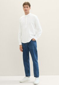 Weißes Langarmhemd mit Stehkragen und Knopfleiste, kombiniert mit blauen gerade geschnittenen Jeans und weißen Sneakern.