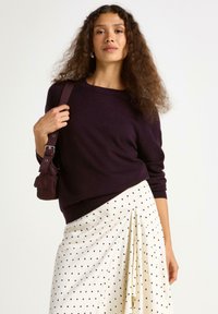 Donkerpaarse trui met lange mouwen, gecombineerd met een crèmekleurige rok met zwarte stippen. Houdt een kleine bruine tas met een hengsel vast.