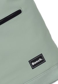 Sac vert clair, à texture lisse, avec une poche zippée à l'avant et une étiquette rectangulaire noire portant le nom de la marque "Bench." en blanc.