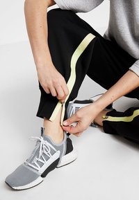 Zwarte sportbroeken met gele zijstrepen en ritsen aan de zoom, gedragen met grijze en witte sneakers. Handen die de rits van de broek aanpassen.