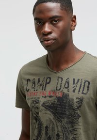 Olive green V-neck t-shirt made of soft cotton; features black and red graphic text and design with road imagery and handwritten-style accents.