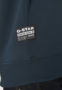 Sweatshirt bleu marine avec un ourlet côtelé et une étiquette noire avec un texte blanc indiquant "G-STAR ORIGINALS RAW DENIM" cousue sur le côté.