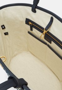 Black leather tote bag interior with beige canvas lining, featuring zippered and slip pockets, gold hardware accents, and brand tags.