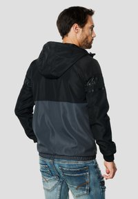 Must ja hall kapuutsiga jope, sileda viimistlusega, millel õlgadel võrgutrikoo paneelid ja soonikuga vöökoht.