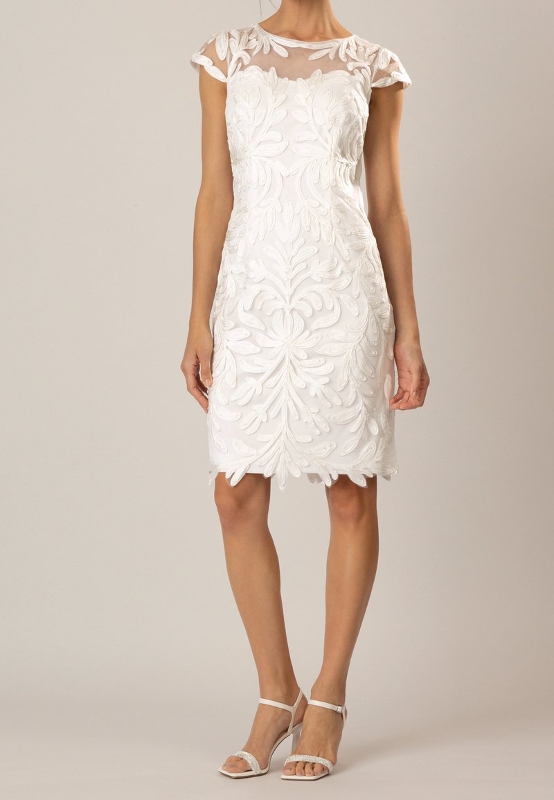 Robe blanche longueur genoux avec motifs floraux brodés, manches courtes transparentes et sandales blanches à talons hauts sur une femme debout.