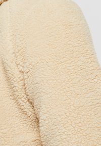 Gros plan sur un tissu en polaire beige avec une surface texturée, présentant une apparence douce et duveteuse et un bord légèrement courbé.