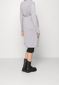 Graues langärmeliges Kleid mit Bindegürtel, seitlichen Schlitzen und weicher Textur, kombiniert mit schwarzen Stiefeletten mit dekorativer Metallverzierung.