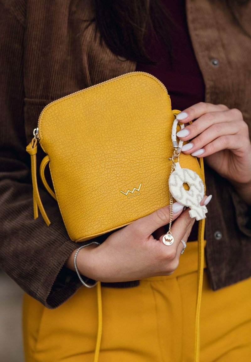 Žlutá crossbody taška s texturou, zapínáním na zip a vnější střapcem. Obsahuje dekorativní klíčenku a logo na přední straně.