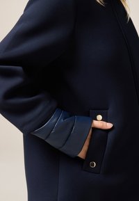 Cappotto navy con un tessuto liscio, dotato di un polsino blu lucido e di una tasca con un dettaglio di bottone a pressione dorato. Design elegante con accenti minimi.