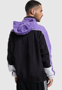 Lahka vetrovka z vijoličasto kapuco in črnim telesom v kombinaciji z belimi detajli. Elastični manšeti in spodnji del, gladka tekstura blaga.