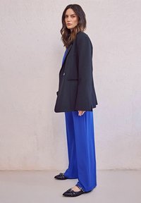 Black tailored blazer over a blue jumpsuit, wide-leg pants, and black flats with metal accents. Smooth fabric, minimalist design.