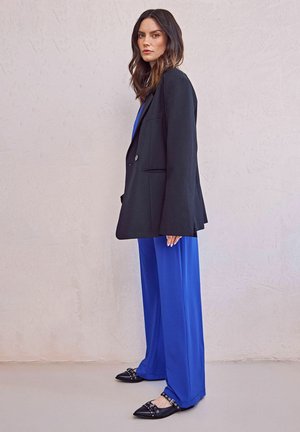 Blazer noir ajusté sur une combinaison bleue, pantalon large, et ballerines noires avec des accents métalliques. Tissu lisse, design minimaliste.