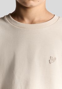 Beige kortærmet t-shirt med ribbet rund hals og et tekstureret, broderet sommerfugl logo på venstre bryst. Glat stof.
